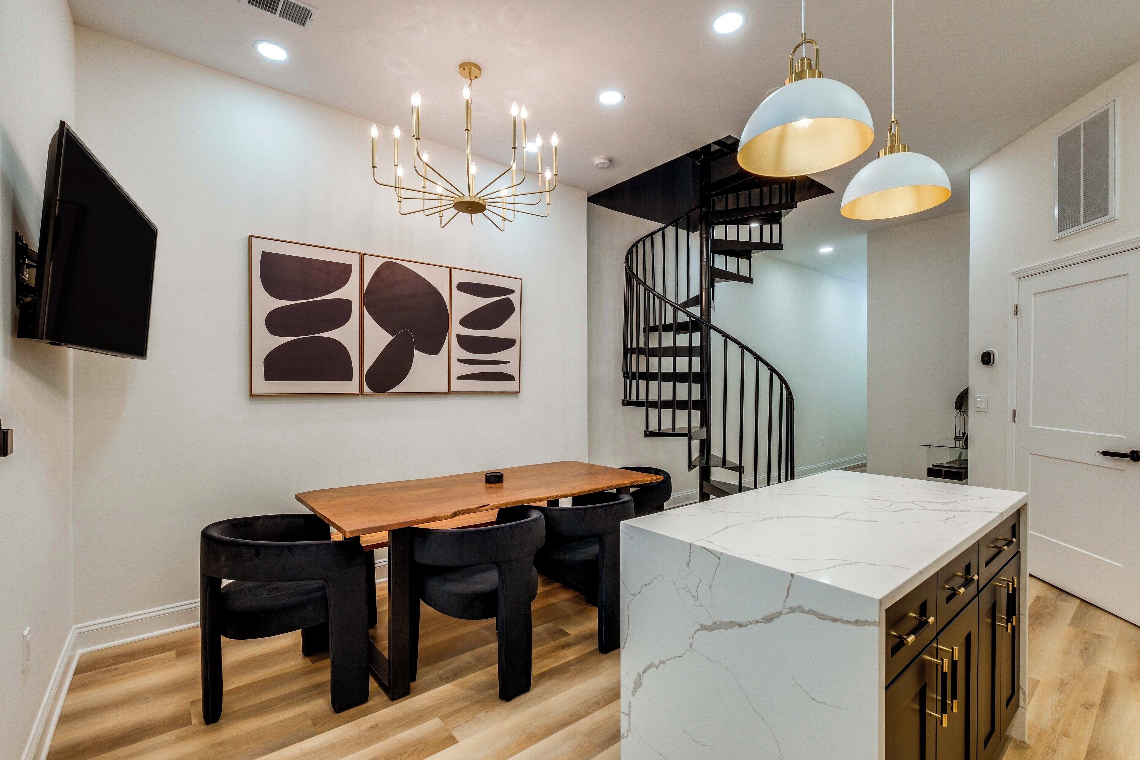 Une cuisine moderne avec une table à manger en bois, des chaises noires, un îlot en marbre, des luminaires suspendus et un escalier en colimaçon noir.