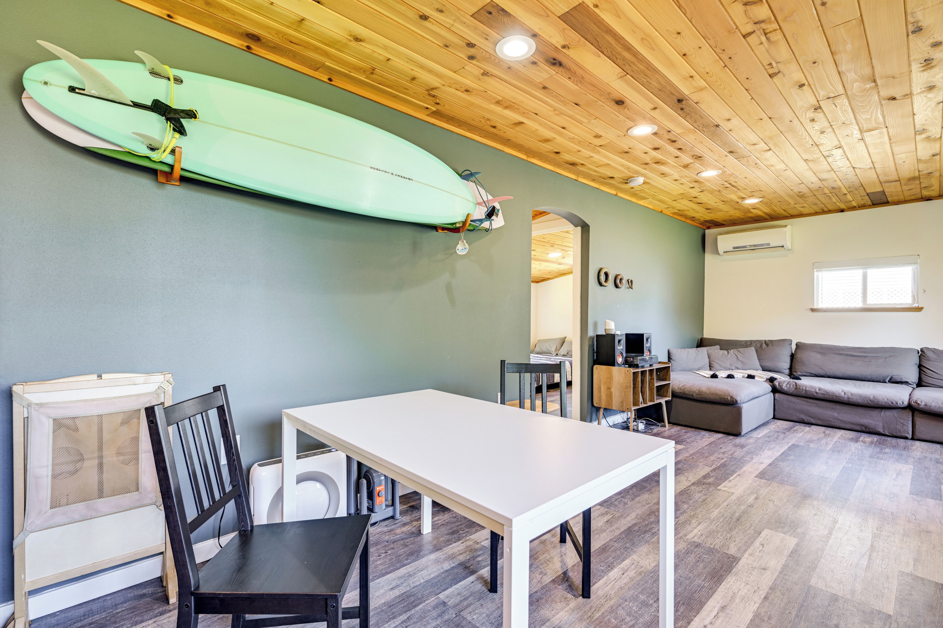 A living room with a wooden ceiling, a white table, a grey sofa, and a surfboard mounted on the wall.