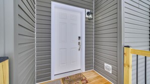 A gray exterior wall with a white door, a small porch with a wooden railing, and a welcome mat. - Walk to Beach: Modern Retreat in Presque Isle! (Presque Isle)