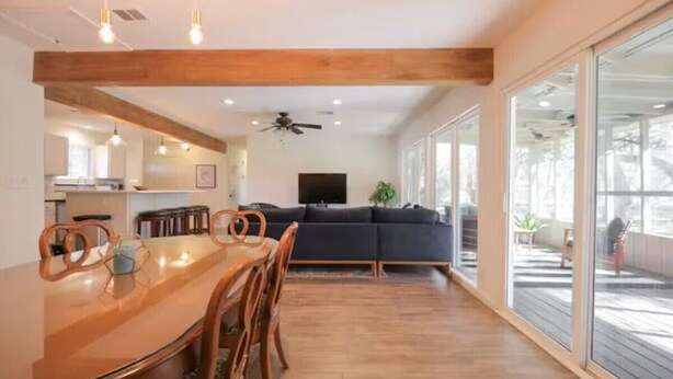 A modern kitchen with wooden beams, a dining area with a glass table, and a living room with a dark sofa and large windows.