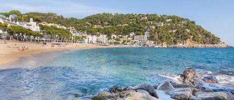 A beach with clear water, rocky shoreline, and buildings on a hillside.