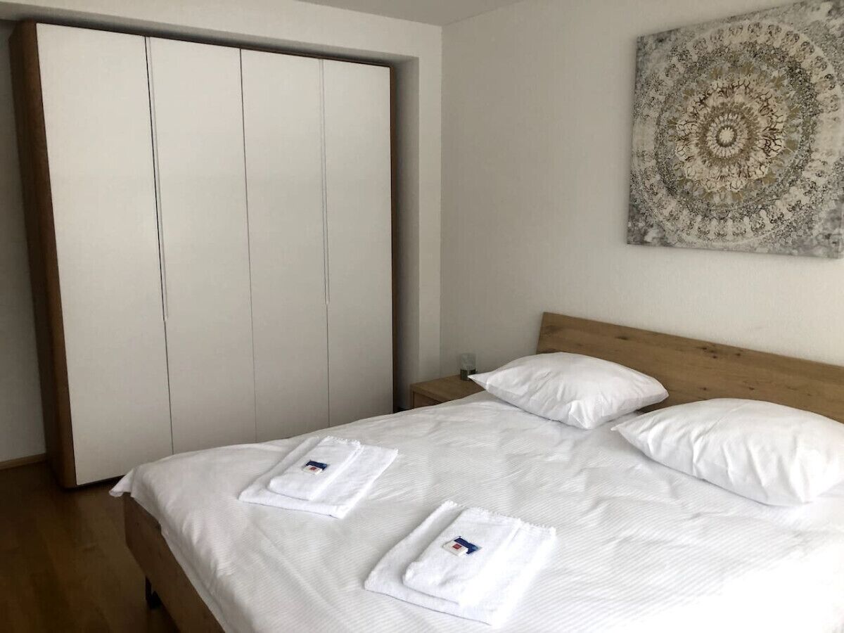 Un lit bien fait, avec du linge de lit blanc et deux serviettes, une tête de lit en bois, une table de chevet avec un verre, et une grande œuvre d’art circulaire accrochée au mur.