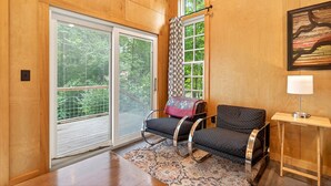 Una habitación con un amplio ventanal corredero, una cortina estampada, un sofá negro con un cojín decorado, una mesita de noche con una lámpara, y un cuadro en la pared.