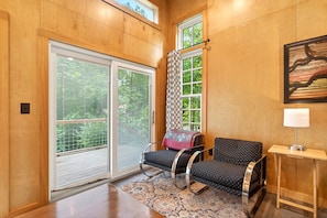 A room with a large sliding glass door, a patterned curtain, a black sofa with a patterned cushion, a small table with a lamp, and a painting on the wall.
