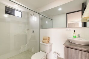 A modern bathroom with a glass shower enclosure, a toilet, a sink with a mirror above it, and a towel placed on the toilet tank. - Torre San Tomas (Medellín)