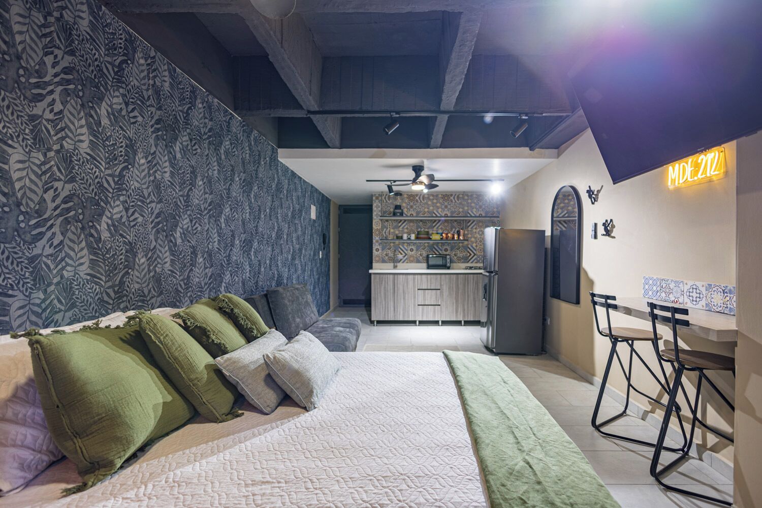 Een moderne slaapkamer met een groot bed, een kitchenette op de achtergrond, en een neonbord aan de muur.