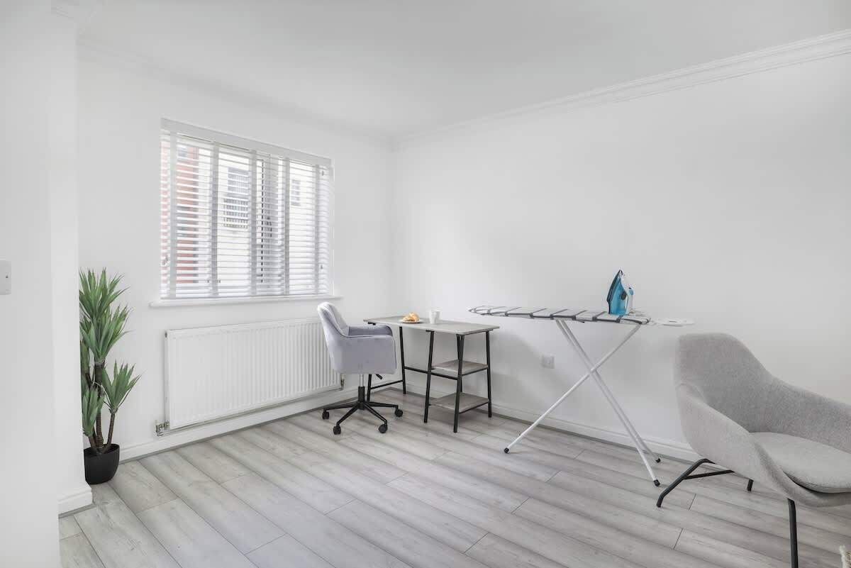 A room with a grey office chair, a small table, a radiator, a window with blinds, and a potted plant.