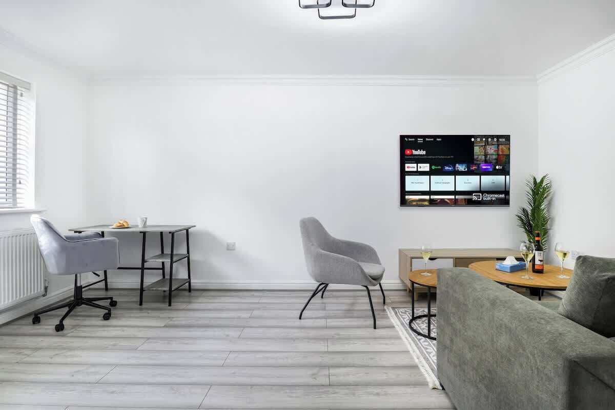 A modern living room with a flat-screen TV mounted on the wall, a grey sofa, a dining table with chairs, and a small plant on the side table.