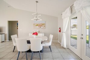 Un comedor con una mesa y sillas blancas, una lámpara colgante y un cuadro con una escena de playa.