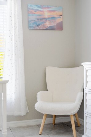 Una habitación con una silla blanca, un tocador blanco y un cuadro de una puesta de sol en la pared.