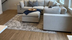 A modern living room with a grey sectional sofa, a small ottoman, and a large painting of a mountainous landscape.