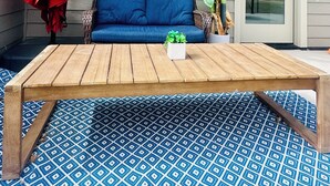 A patio with a wooden table, blue patterned rug, a blue sofa, a red umbrella, and a potted plant.