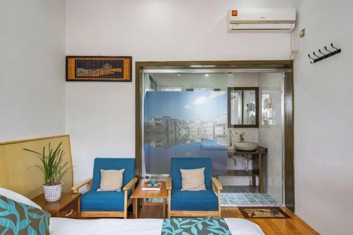 A hotel room with a bed, two blue armchairs, a small table, a potted plant, a framed picture, a bathroom with a sink and mirror, and an air conditioning unit.