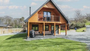A two-story wooden cabin with a balcony, a covered porch, and a gravel driveway. - Sunset Lodge 5 (Llanbedr)