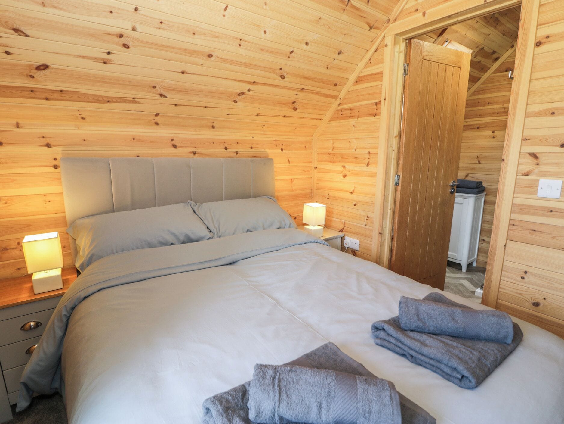 A cozy wooden cabin bedroom with a bed, bedside lamps, and neatly folded towels.