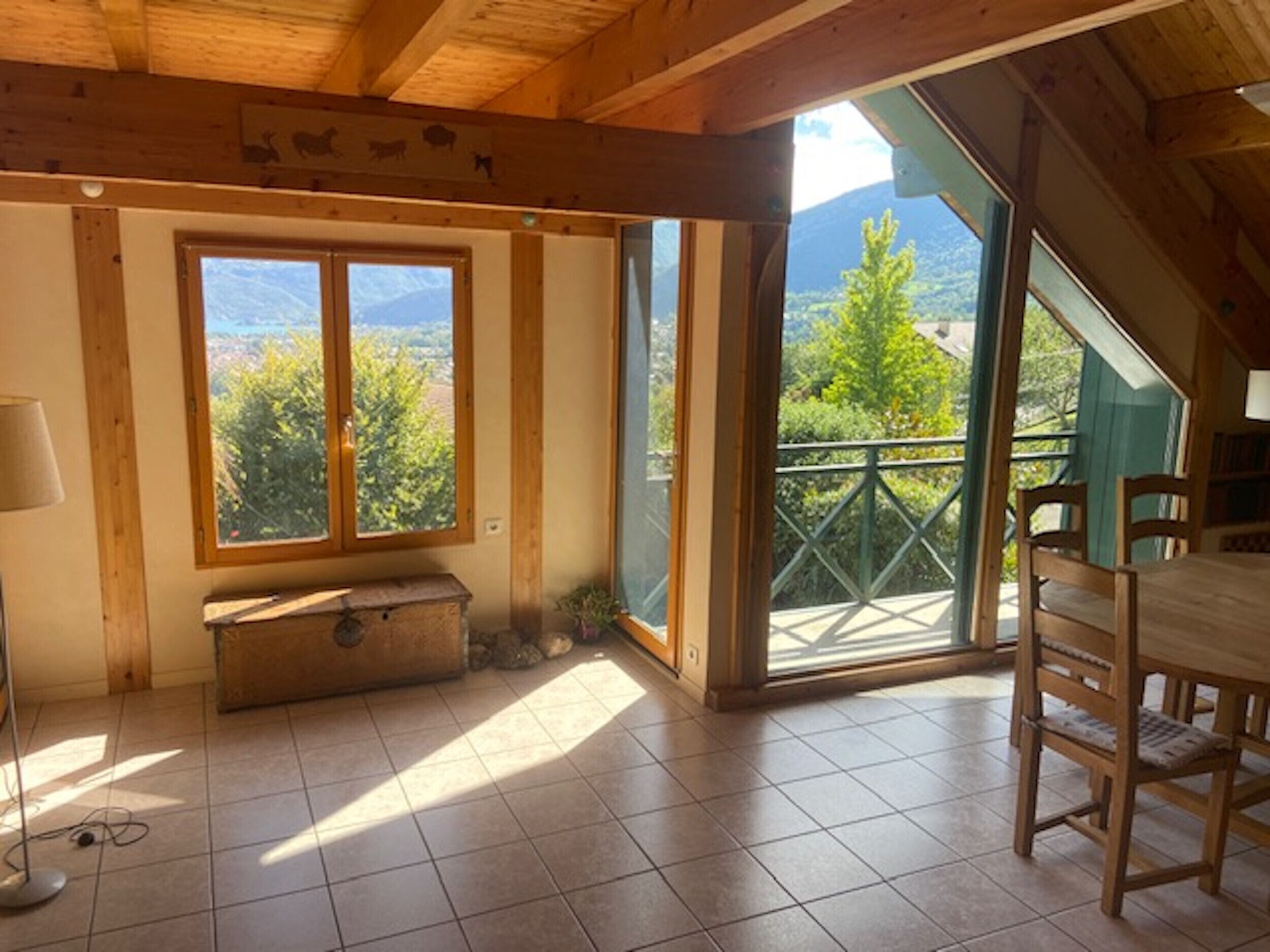 A room with a wooden ceiling, a large window with a view of trees and mountains, a wooden bench, and a dining table with chairs.