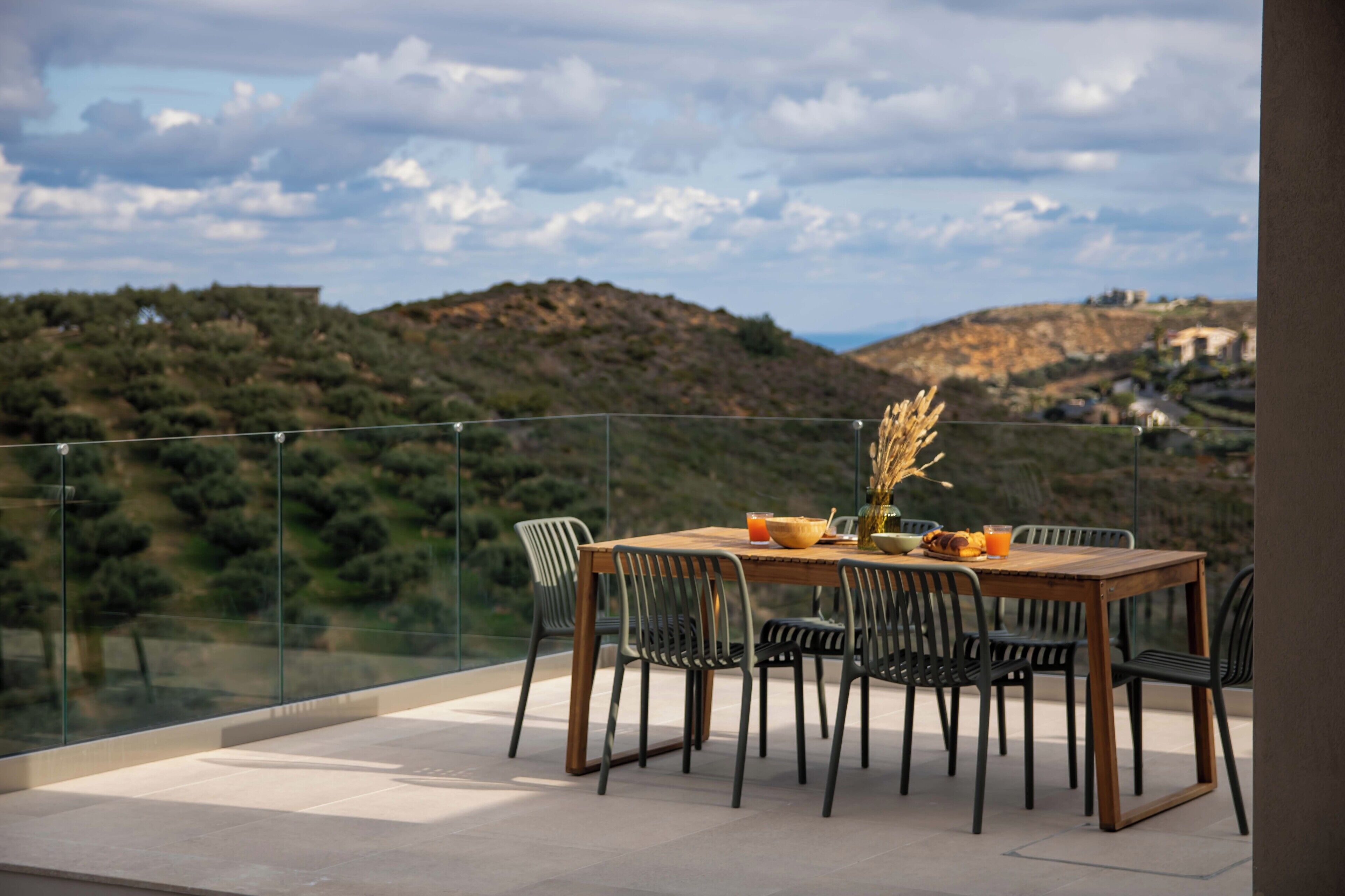 A dining table with chairs on a balcony with a glass railing, overlooking a hilly landscape.