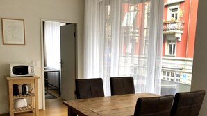 Un comedor con una mesa de madera y cuatro sillas, un microondas sobre una pequeña estantería, y vista a un edificio rojo a través de cortinas traslúcidas.