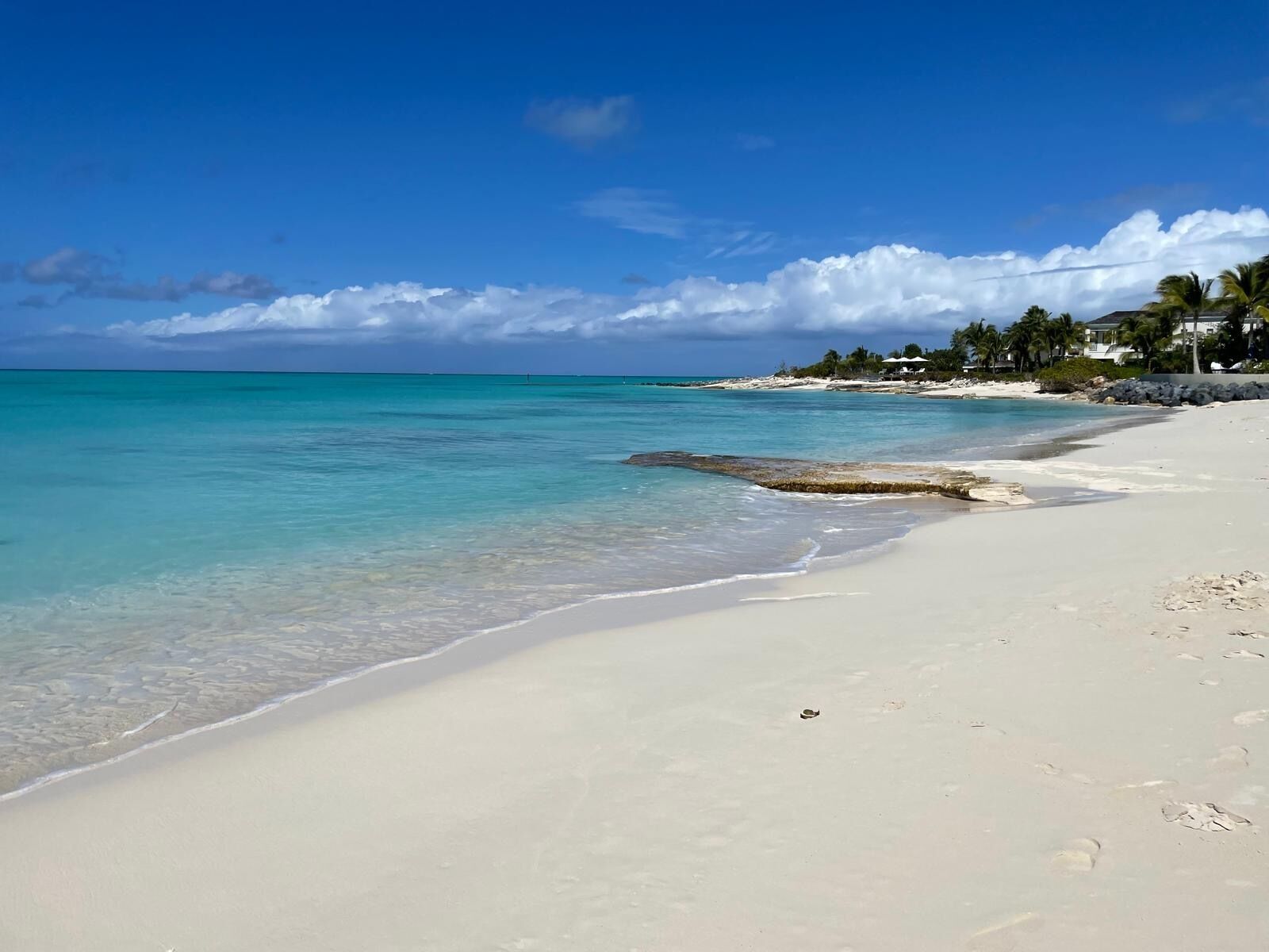 A beach with clear turquoise water, a sandy shore, and a distant shoreline with buildings and palm trees.