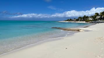 Una playa con agua turquesa cristalina, una orilla arenosa y una costa distante con edificios y palmeras.
