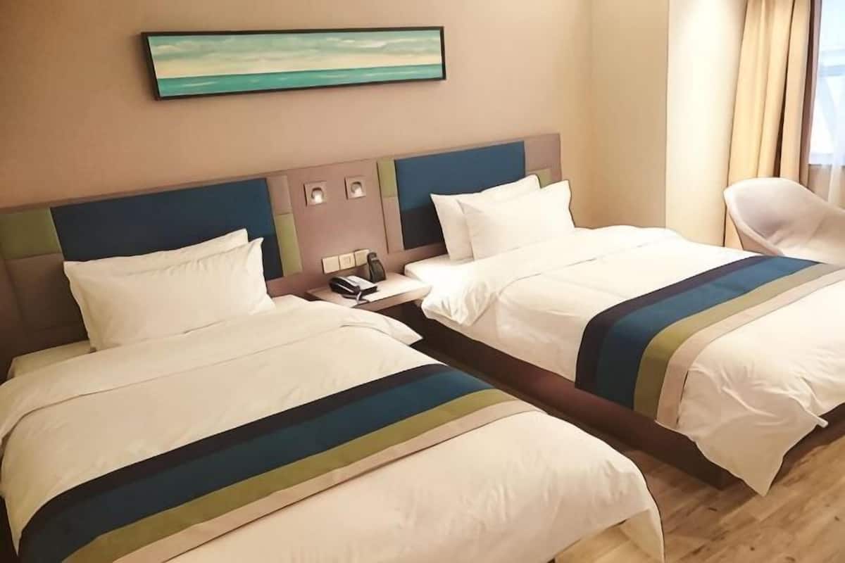 A hotel room with two beds, a nightstand, a painting on the wall, and a window with curtains.