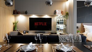 En moderne stue med et stort fladskærms-tv, der viser 'NETFLIX', et spiseområde med dækkede borde og en bar med glas.