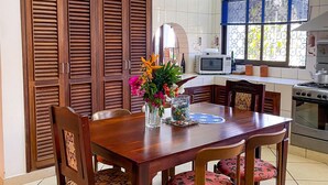 A dining area with a wooden table and chairs, a vase with flowers, and a kitchen in the background. - Villa Sublime- Diani Beach 4 Bedrooms 50 meters from the beach- Luxurious🌴🔥⛱️ (Diani Beach)