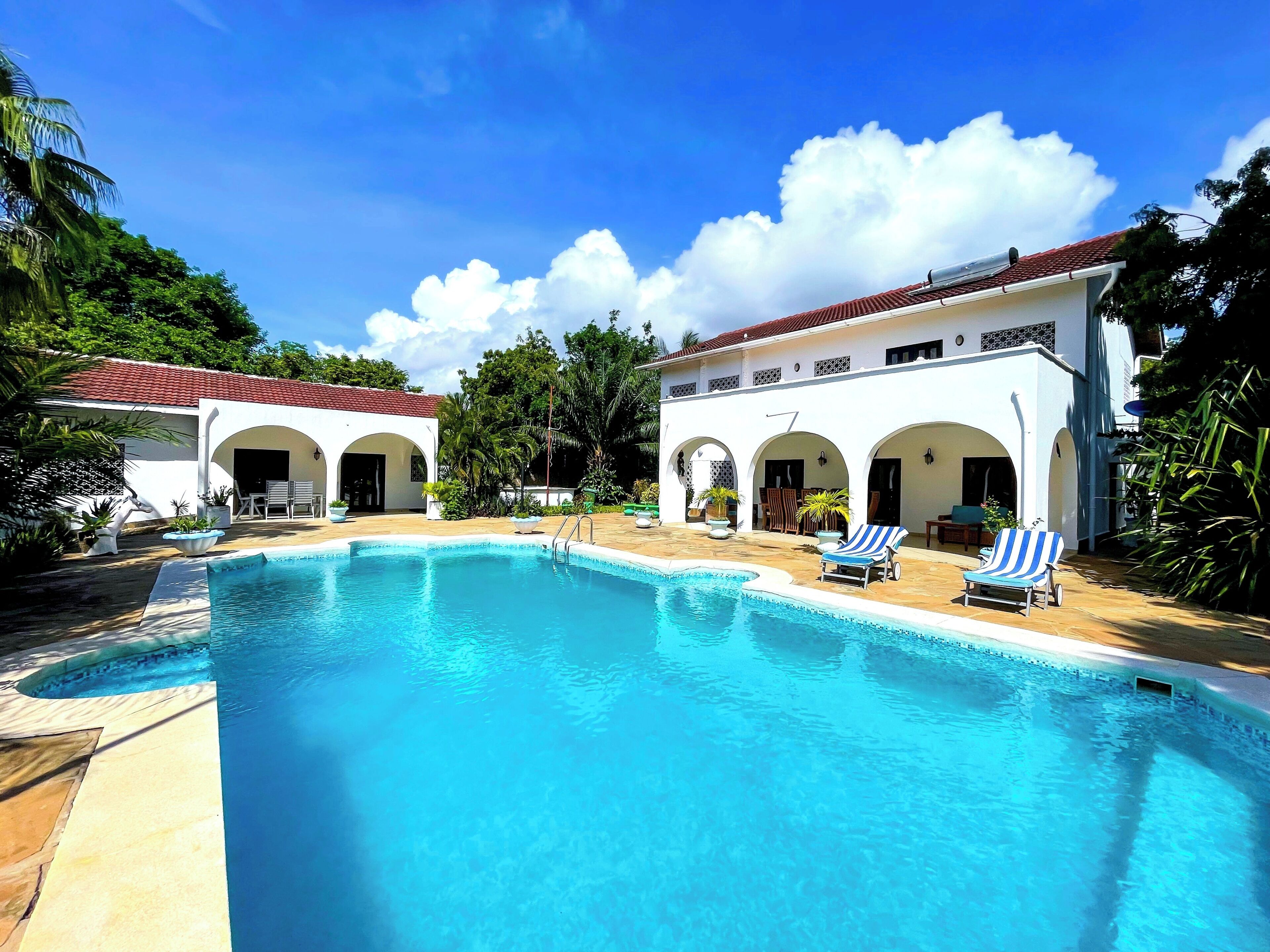 A swimming pool with lounge chairs, a white building with arches, and surrounding greenery.