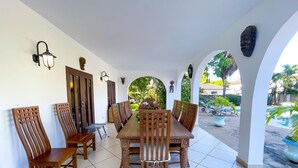 A covered dining area with a long wooden table and chairs, surrounded by white walls with decorative wall sconces and a view of a garden and pool. - Villa Sublime- Diani Beach 4 Bedrooms 50 meters from the beach- Luxurious🌴🔥⛱️ (Diani Beach)