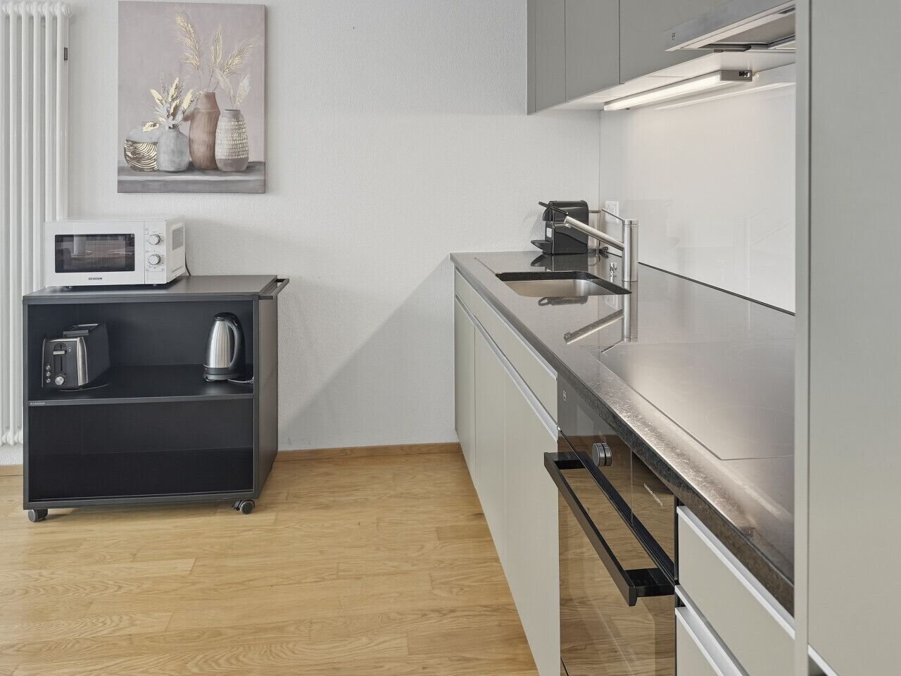 A modern kitchen with a microwave, toaster, and kettle on a black cart, a stainless steel sink, and a painting on the wall.