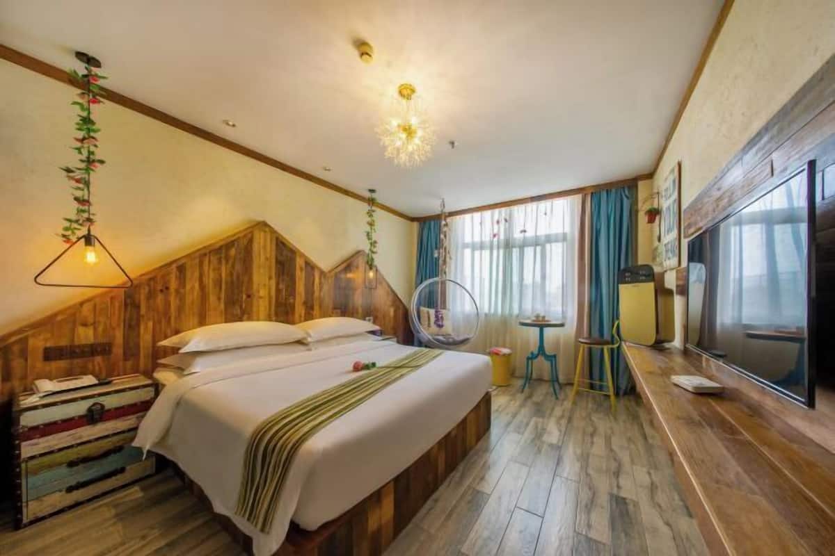 A bedroom with a wooden headboard, a bed with white linens, a hanging light fixture, a round chair, a small table, and a TV on the wall.