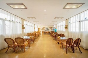 A spacious dining area with long tables and wicker chairs, large windows on both sides, and a kitchen in the background. - Shuiyue Qinghua Hotel (Deqing)