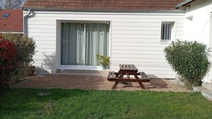 Une maison de plain-pied avec un toit en tuiles, une grande porte-fenêtre coulissante et une petite fenêtre. On trouve une table de pique-nique en bois dans le jardin.
