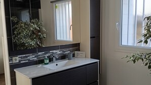 Une salle de bain moderne avec un grand miroir, des meubles de couleur sombre, un lavabo blanc et une fenêtre équipée de stores.