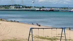 A beach with a swing set in the foreground, a pier with a water tower in the background, and a bridge across the water. - Lake Cottage at Beachcomber of Mackinaw (Mackinaw City)