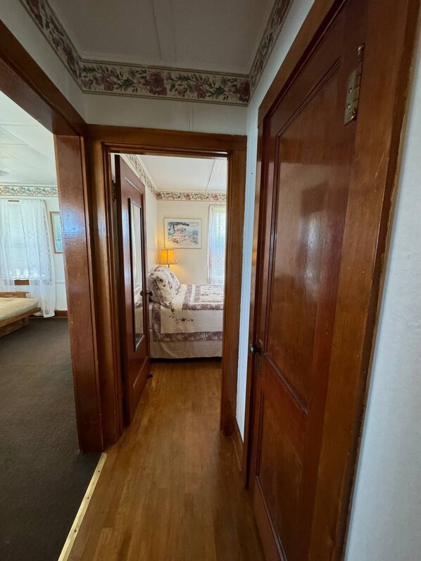 A hallway leading to a bedroom with a bed, a lamp, and a painting on the wall. - Lake Cottage at Beachcomber of Mackinaw (Mackinaw City)