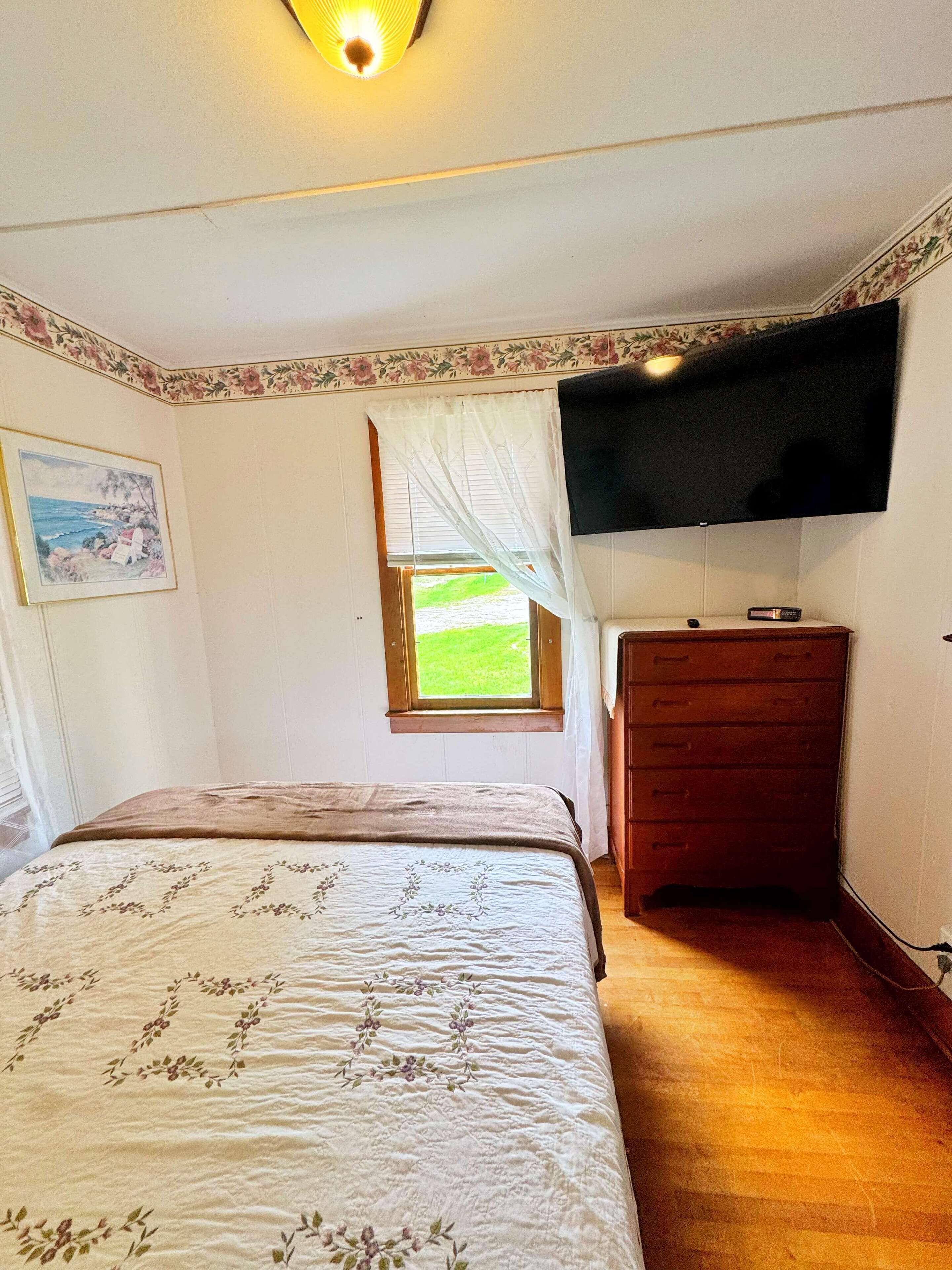 Quarto com cama, cômoda, televisão e janela com vista para o exterior.