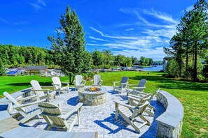 Un espace extérieur circulaire aménagé avec des chaises Adirondack et un foyer, entouré de murs en pierre, le tout offrant une vue sur un lac et une forêt en arrière-plan.