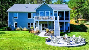 Une maison à deux étages dotée d’un porche à l’avant, d’une terrasse et d’un espace extérieur circulaire aménagé avec des chaises et un foyer.
