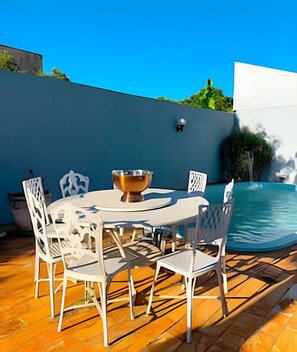 Una mesa redonda con un bol central, rodeada de sillas blancas, sobre una terraza de madera con una piscina al fondo.