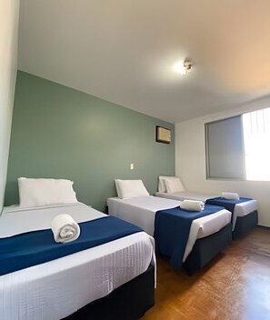 Habitación de hotel con tres camas, cada una con una almohada blanca y una toalla enrollada. La habitación tiene una pared verde, una ventana y un suelo de madera.