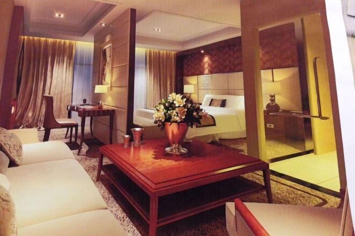 A hotel room with a sofa, a wooden coffee table with a vase, a desk, a chair, a bed, and a glass partition.