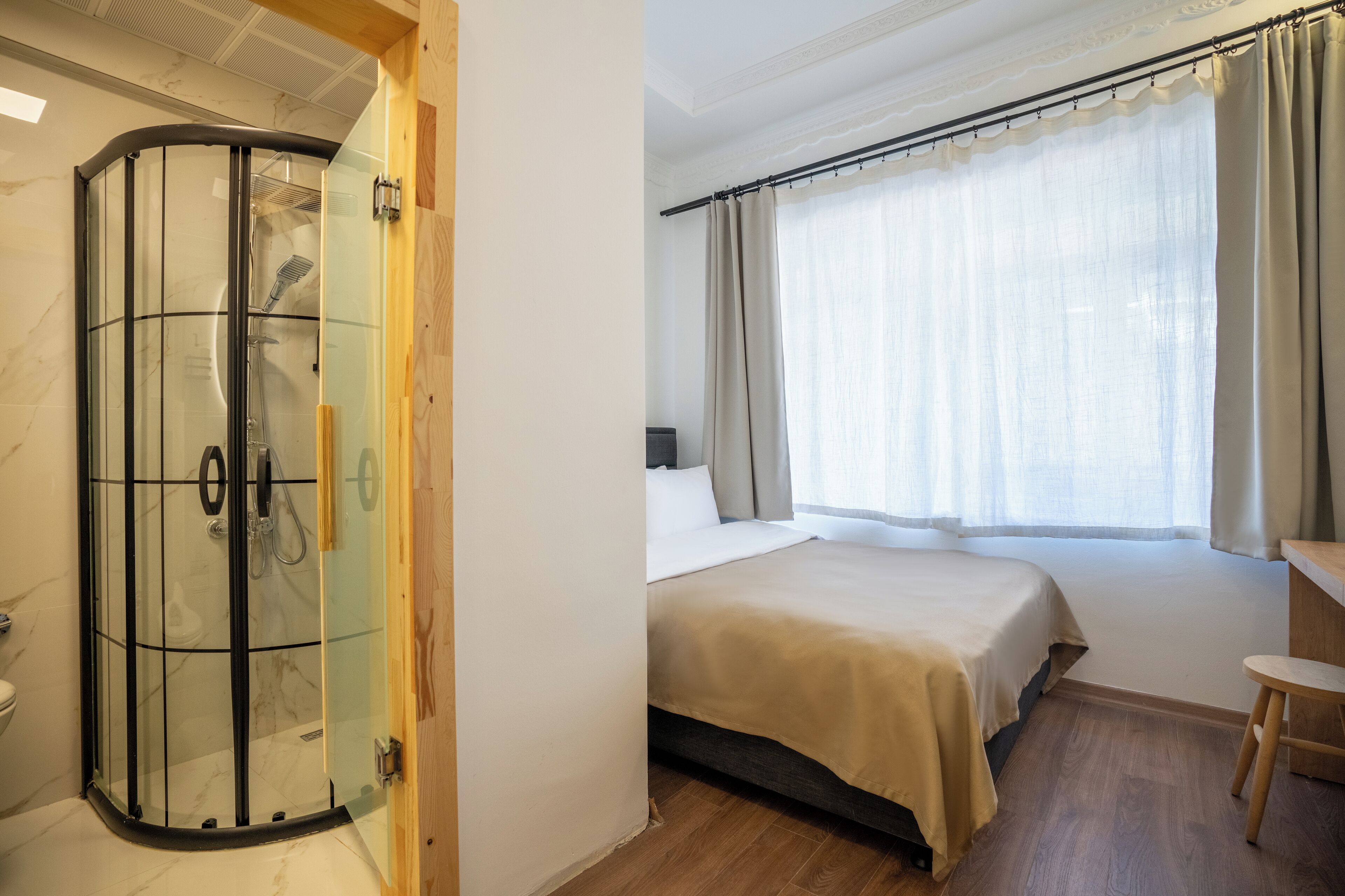 A hotel room with a glass-enclosed shower, a bed with a wooden headboard, a wooden desk, and a window with curtains.