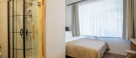 Habitación de hotel con ducha envuelta en vidrio, cama con cabecera de madera, escritorio de madera y ventana con cortinas.