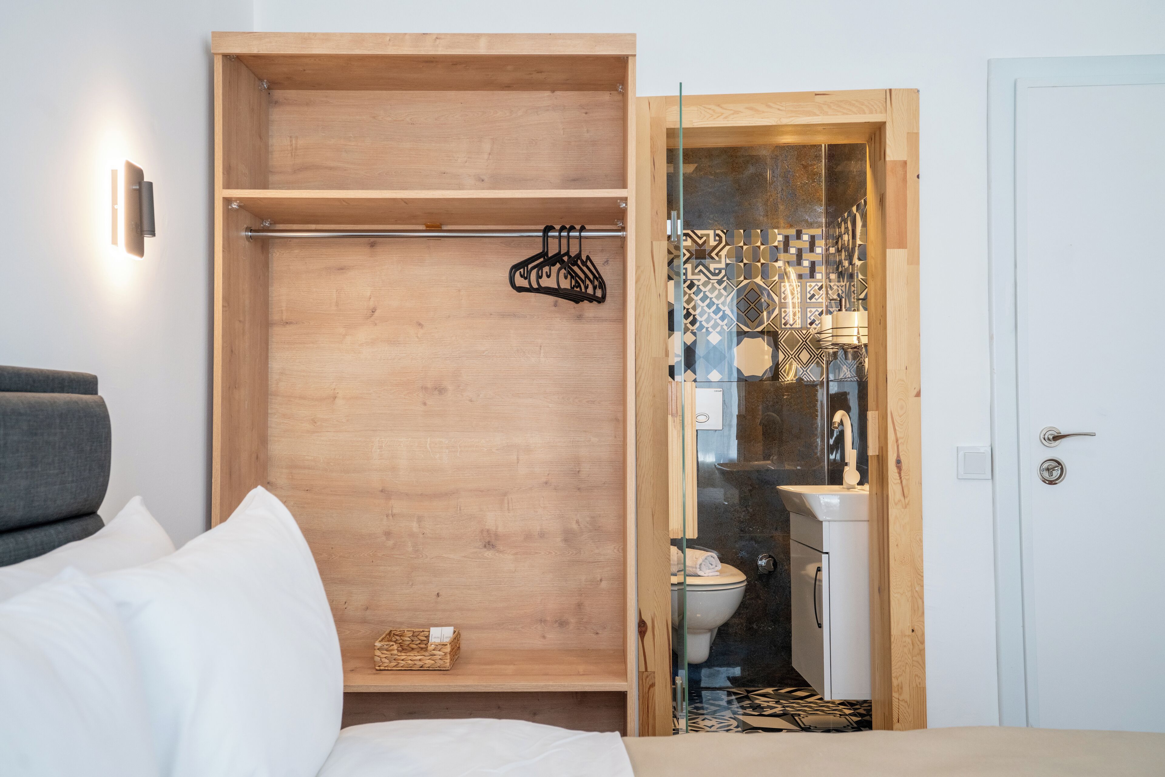 Un dormitorio compacto con un armario de madera, un baño con mampara de vidrio y un sofá gris.