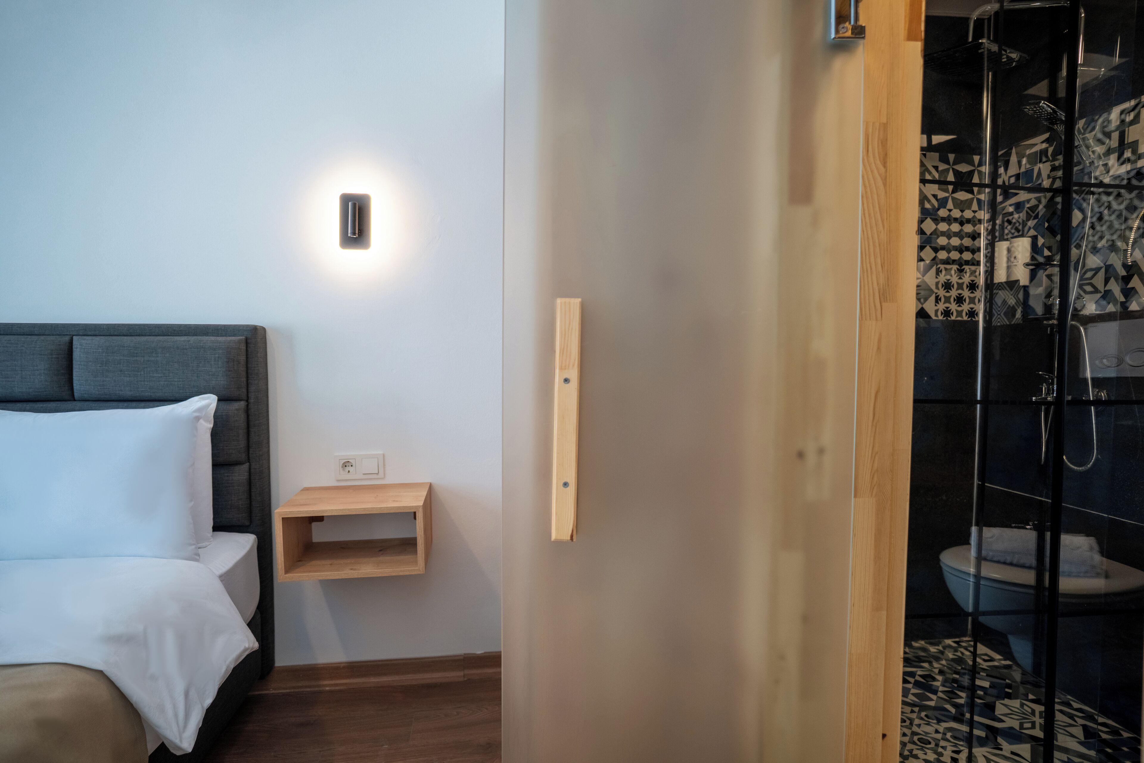 Una camera d'albergo moderna con un letto, un comodino, una lampada a parete e uno spazio doccia.