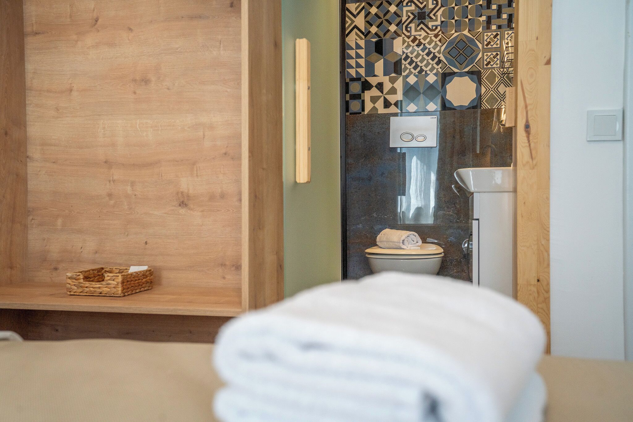Un baño con tocador de madera, inodoro con salpicadero de azulejos estampados y una toalla en el suelo.