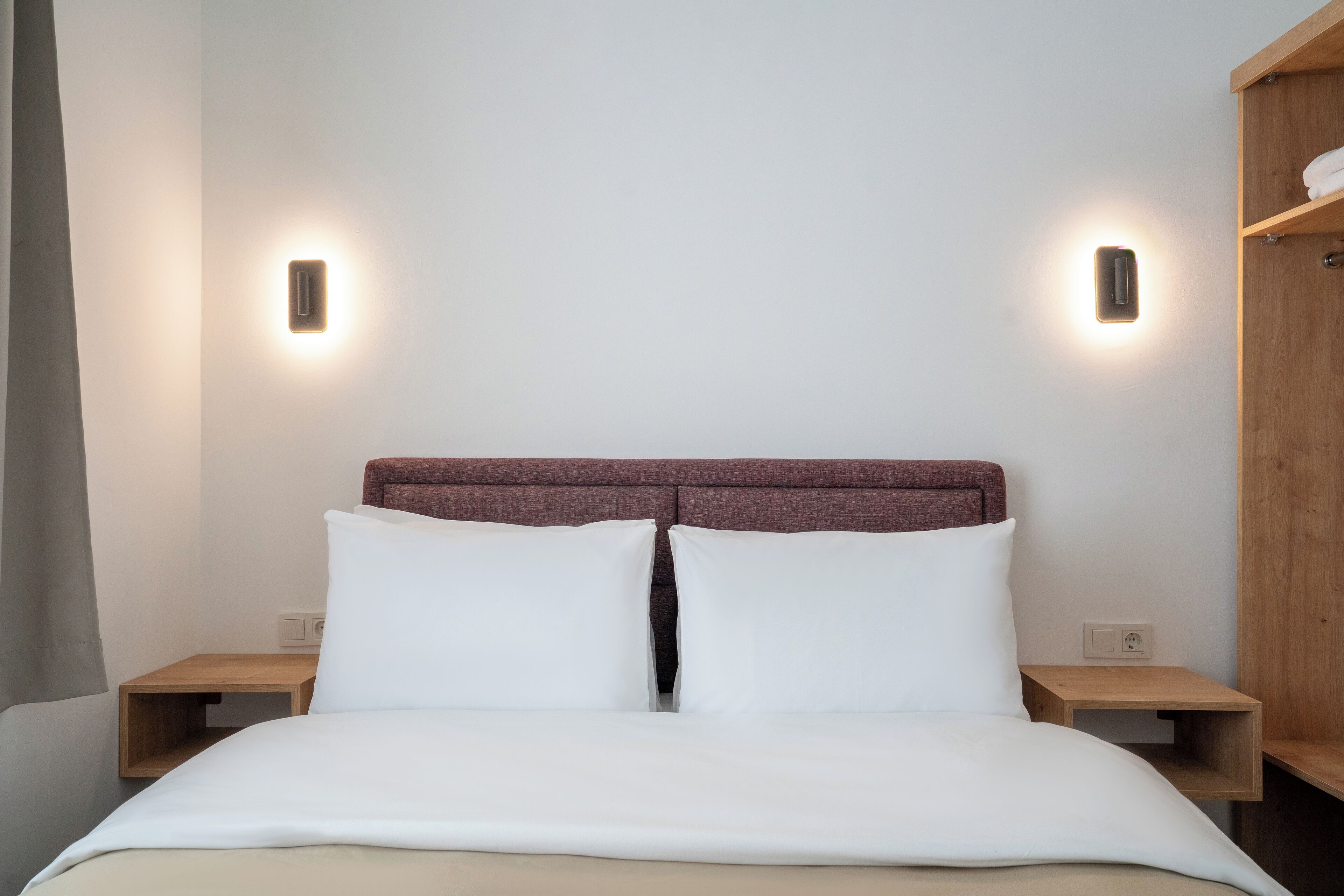 A neatly made bed with white linens, two bedside tables with wooden nightstands, and a headboard with a reddish-brown panel.