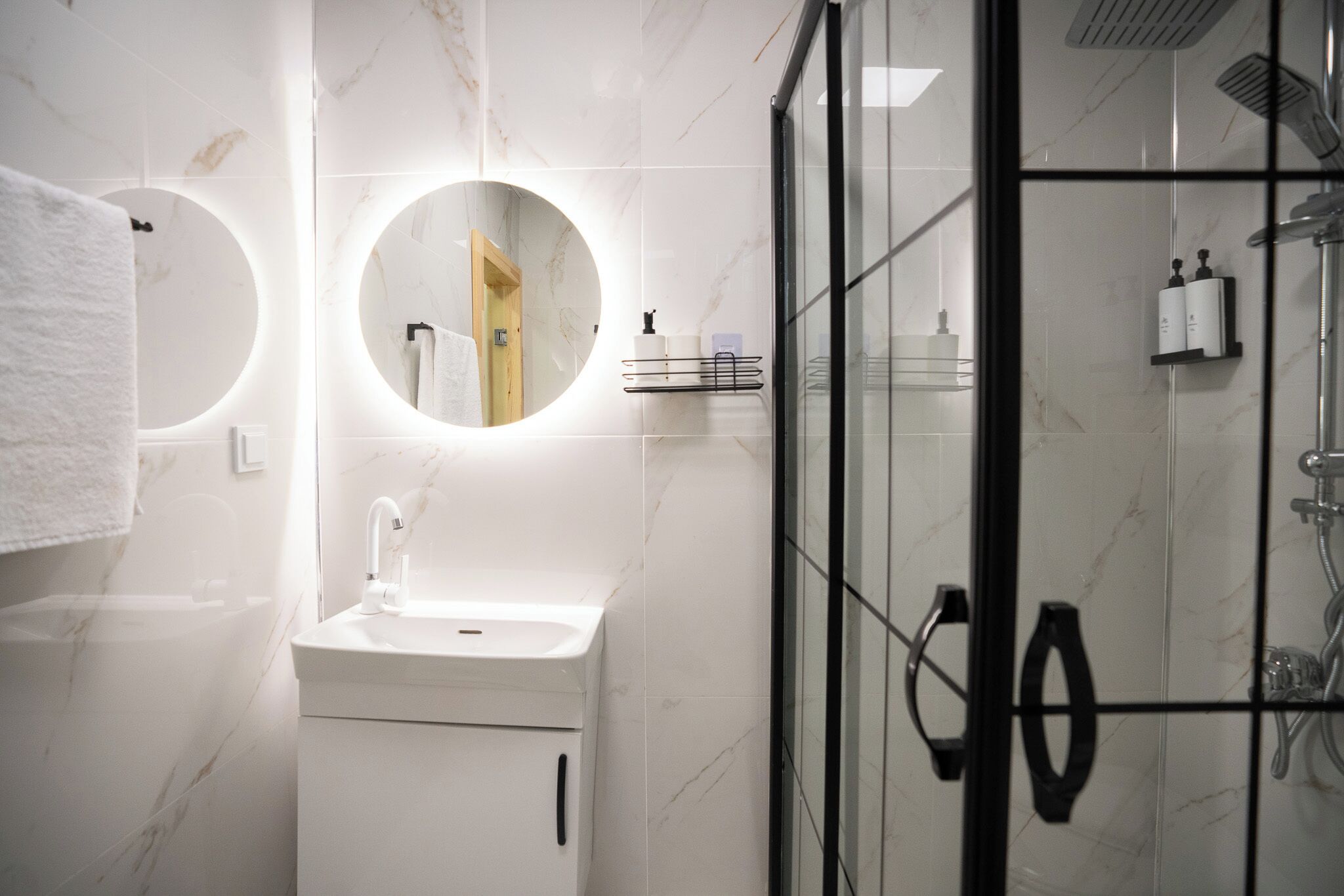 Une salle de bain moderne avec un lavabo blanc, un miroir rond et une douche séparée par des parois en verre.