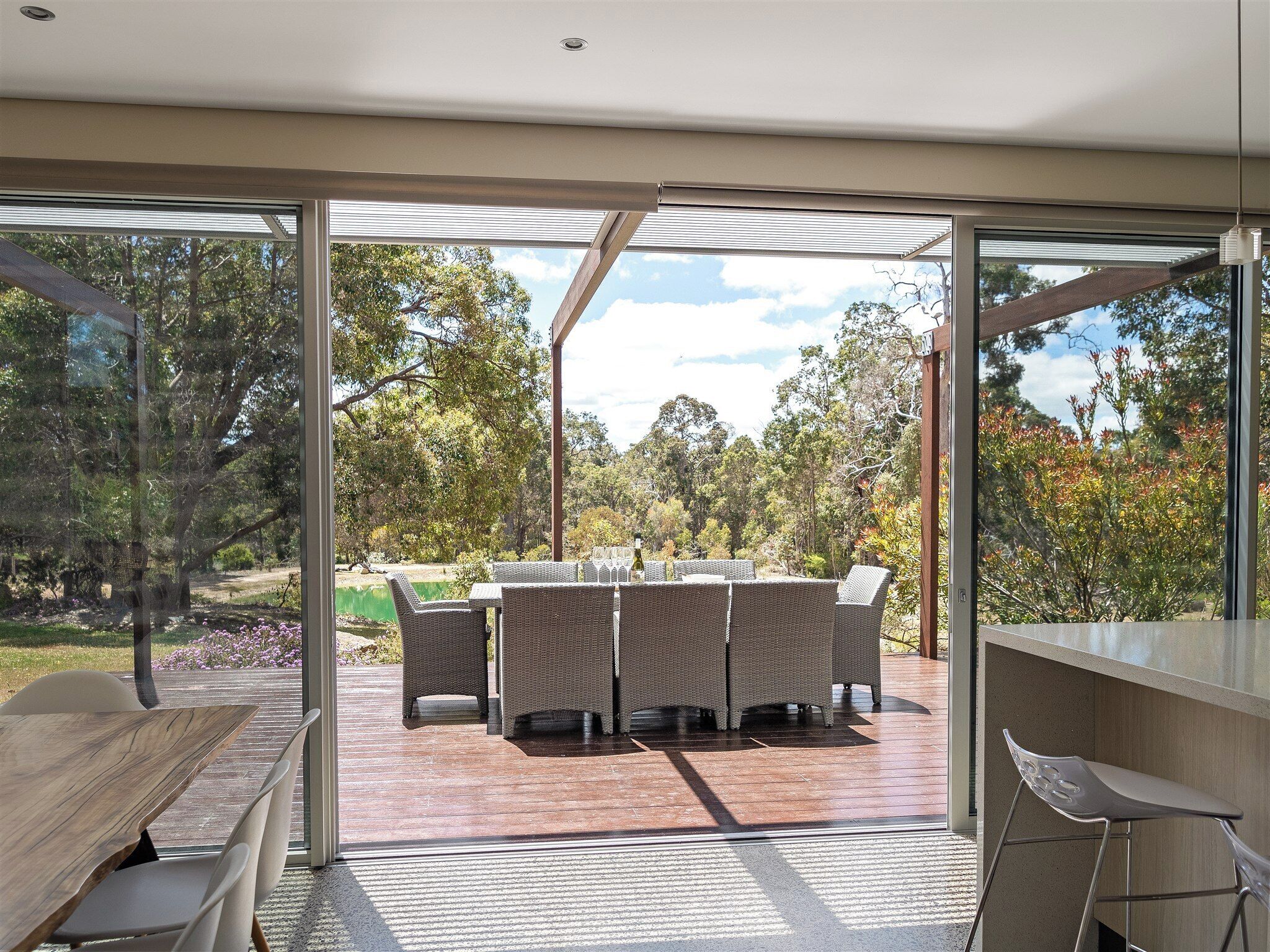 A modern outdoor dining area with a glass-enclosed patio, wicker furniture, and a view of a natural landscape.